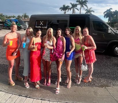 Eight women in colorful cocktail dresses holding drinks and posing in front of a black party shuttle with palm trees and a sunny coastal Florida backdrop.
