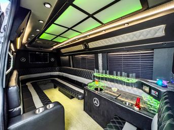 LED-lit luxury party van interior with green ceiling panels, quilted black-and-gray leather wraparound seating, mini bar with glass holders and cupholders, and wood-look flooring