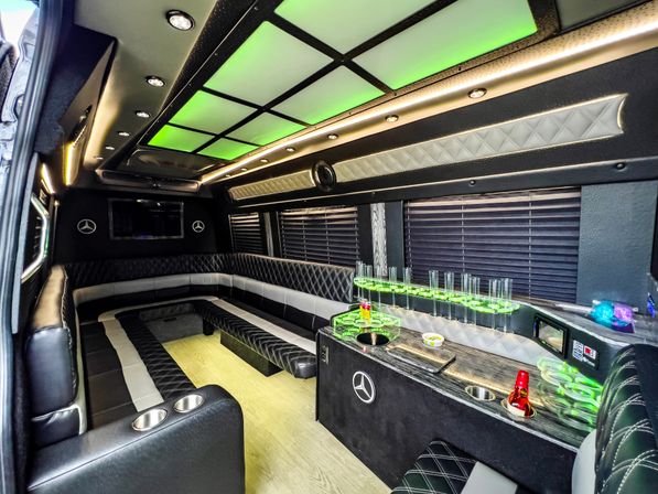 LED-lit luxury party van interior with green ceiling panels, quilted black-and-gray leather wraparound seating, mini bar with glass holders and cupholders, and wood-look flooring