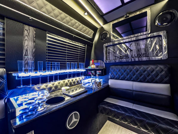 LED-lit luxury party van interior with quilted black leather bench seating, blue ambient lighting, illuminated stemware rack and mini bar with bottles, wood-grain accents, speakers and wall-mounted screen — modern VIP shuttle vibe.