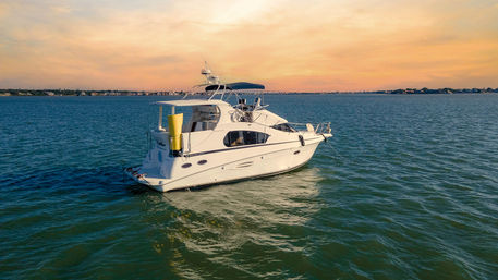 Sleek white motor yacht anchored in a calm blue-green bay at sunset, golden sky and distant coastal shoreline visible.