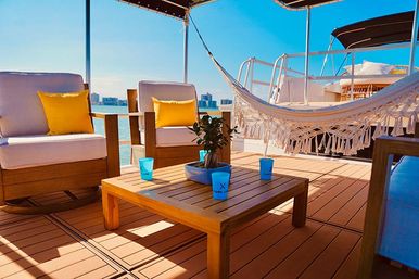Bright yacht deck lounge with wooden chairs and yellow pillows, macramé hammock, low coffee table with blue cups and potted plant, turquoise water and distant skyline