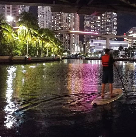 Night paddleboarder gliding on an urban waterfront canal with palm trees and high-rise buildings casting colorful reflections.