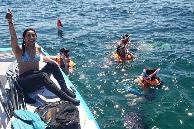 Smiling woman on a small boat gives a thumbs-up as three snorkelers in orange life jackets, masks and fins float in clear blue ocean water on a sunny day