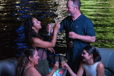 Four people smiling and toasting with champagne on a nighttime boat ride, colorful lights reflecting on the river water.