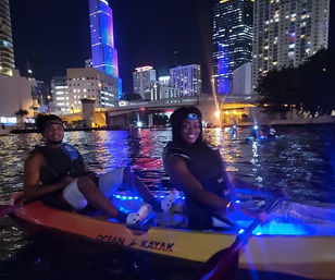 Two people kayaking at night in a blue-lit kayak on an urban waterfront, smiling as illuminated skyscrapers and a bridge reflect on the water.