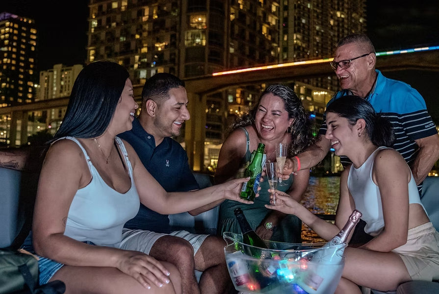 Group of friends laughing and toasting with beer and champagne on a boat at night, with an illuminated city skyline and bridge reflecting on the water.