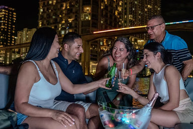 Group of friends laughing and toasting with beer and champagne on a boat at night, with an illuminated city skyline and bridge reflecting on the water.