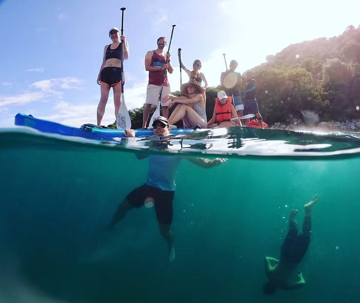 Split-level sunlit shot of a group of paddleboarders on clear turquoise water by a lush tropical shoreline, with one person floating at the surface and another diving underwater