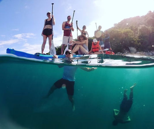 Split-level sunlit shot of a group of paddleboarders on clear turquoise water by a lush tropical shoreline, with one person floating at the surface and another diving underwater