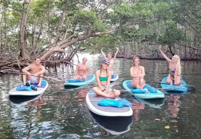 Six smiling people on stand-up paddleboards (SUP) paddling and posing in a calm mangrove-lined tropical creek.