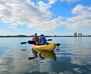 Two smiling paddlers in life jackets in a yellow tandem kayak on calm lake water, reflected clouds and a distant shoreline with waterfront buildings under a bright blue sky.