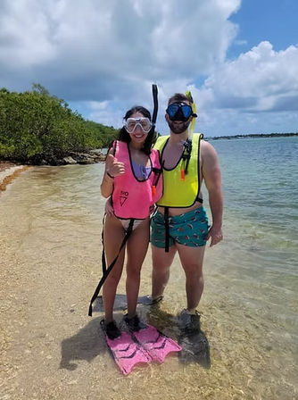 Two snorkelers wearing bright pink and yellow life vests and masks, one giving a thumbs-up, standing on a tropical sandy shoreline with pink fins in shallow clear water, mangroves and a blue sky with fluffy clouds.