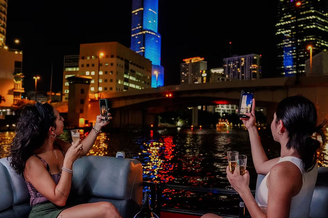 Two people on a lively nighttime river cruise holding champagne and taking smartphone photos of a brightly lit downtown waterfront skyline with colorful skyscraper reflections on the water.