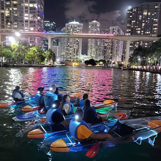 Group of paddlers in clear LED kayaks on a downtown waterfront at night, colorful bridge lights and high-rise skyline reflecting pink, blue and green hues on the water with palm trees along the shore.