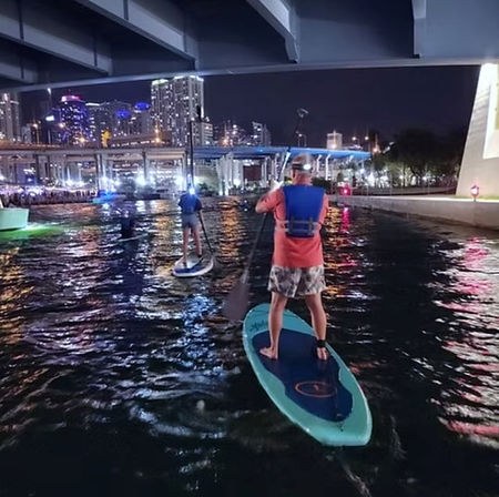 Two stand-up paddleboarders in life vests gliding under a bridge on a lit urban waterway at night, city skyline and colorful lights reflecting on the water.