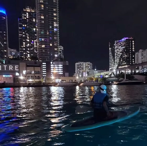 Person paddling an LED-lit paddleboard at night on an urban waterfront, downtown high-rise skyline and marina lights reflecting on rippling water.