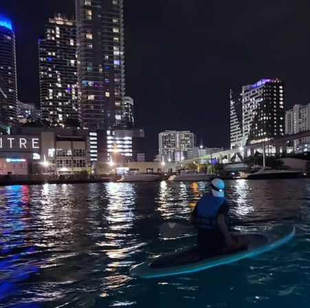 Person paddling an LED-lit paddleboard at night on an urban waterfront, downtown high-rise skyline and marina lights reflecting on rippling water.