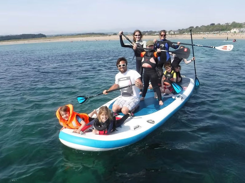 Group of adults and children on an oversized stand-up paddleboard (SUP) in clear coastal ocean water near a sandy beach, kids in life jackets and paddles raised on a sunny day.