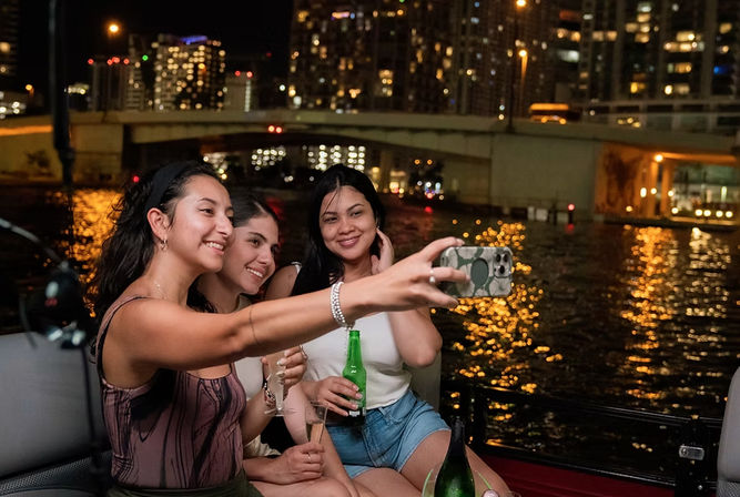 Three friends taking a selfie on a nighttime city boat ride, holding drinks with bridge and city lights reflecting on the water.