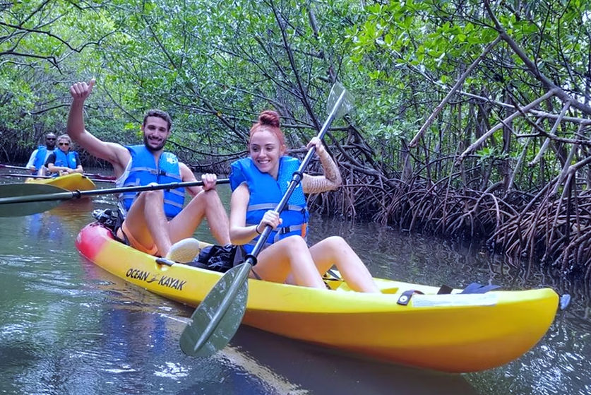 Two smiling paddlers in blue life vests navigating a yellow tandem kayak through a narrow tropical mangrove tunnel with exposed tangled roots
