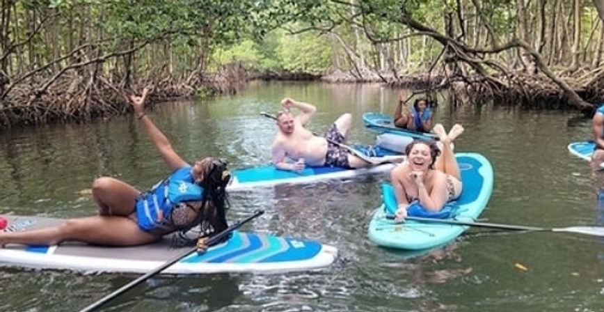 Group of people laughing and lounging on paddleboards in a mangrove-lined coastal creek, wearing life vests and swimwear with paddles — fun paddleboarding outing.