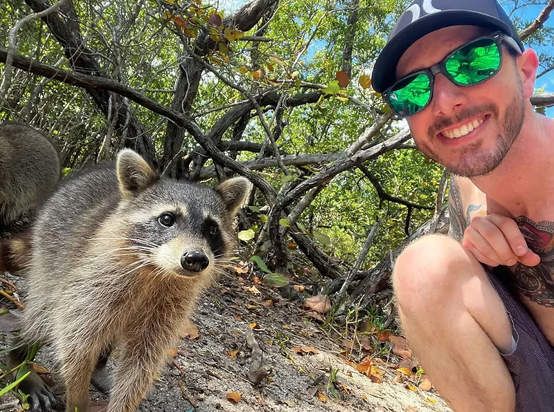 Curious raccoon in sandy coastal mangrove approaches the camera while a smiling man in a cap and green mirrored sunglasses crouches nearby — outdoor wildlife selfie.