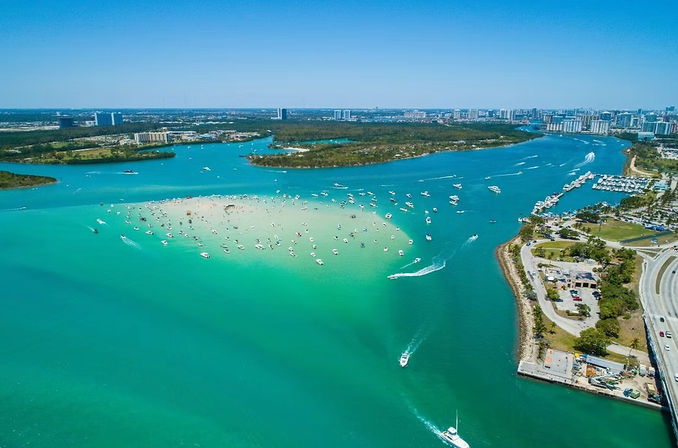 Aerial view of a sunny turquoise bay with a crowded sandbar dotted with dozens of anchored boats and a distant coastal city skyline