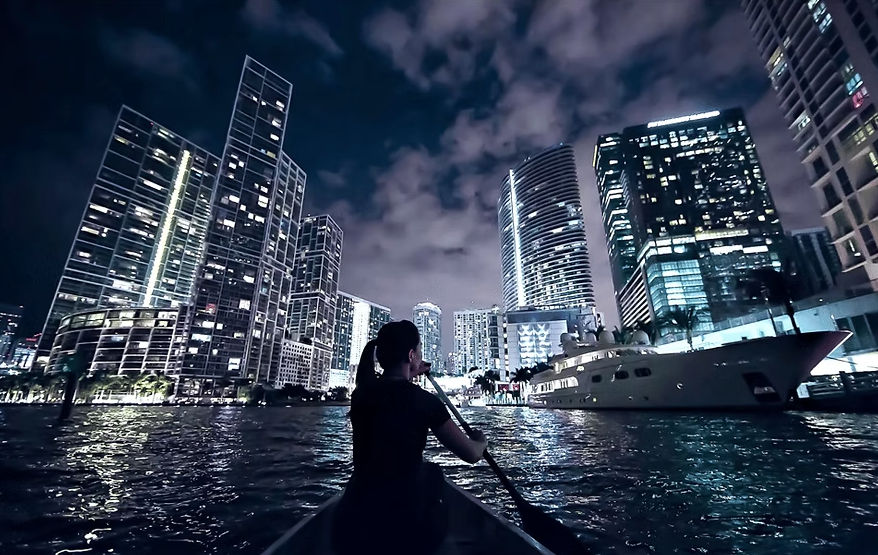 Person paddling a canoe at night toward an illuminated downtown skyline and docked yacht, city lights reflecting on rippling water.