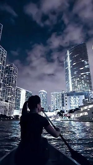 Person paddling a canoe at night toward an illuminated downtown skyline and docked yacht, city lights reflecting on rippling water.