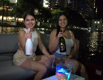 Two friends on a nighttime boat party holding champagne bottles, waterfront city lights reflecting on the water, illuminated ice bucket and flutes.