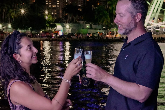 Two people clinking champagne flutes at a city waterfront at night, toasting under illuminated skyline and Ferris wheel with reflections on the water.