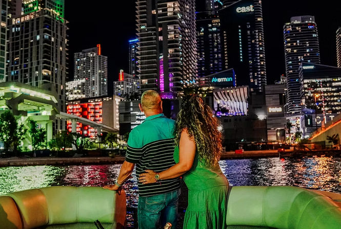 Couple on a boat at night overlooking a neon-lit downtown waterfront skyline with tall illuminated skyscrapers reflecting in the water