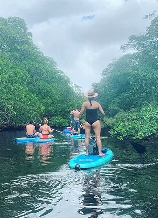 Stand-up paddleboarders navigating a narrow mangrove-lined tropical waterway — woman in a black swimsuit and straw hat standing on a turquoise board in the foreground, others paddling ahead under a cloudy sky.