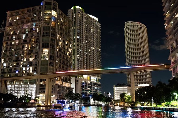 Nighttime Miami riverfront skyline with illuminated high-rise apartments, a rainbow-lit elevated rail bridge and a boat casting colorful reflections on the water