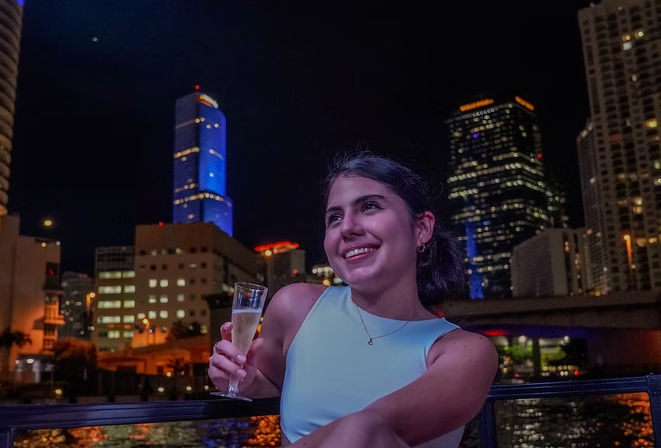 Smiling young woman in a light-blue sleeveless top holding a champagne flute on a waterfront balcony at night, with illuminated downtown skyscrapers and their reflections sparkling on the water behind her.