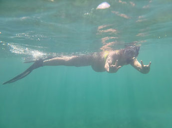 Underwater shot of a swimmer wearing a mermaid-style tail, gliding near the surface in clear turquoise water while flashing double shaka hand signs.