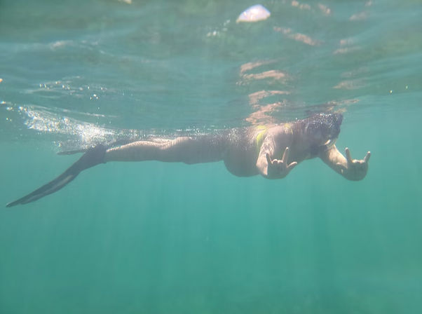 Underwater shot of a swimmer wearing a mermaid-style tail, gliding near the surface in clear turquoise water while flashing double shaka hand signs.