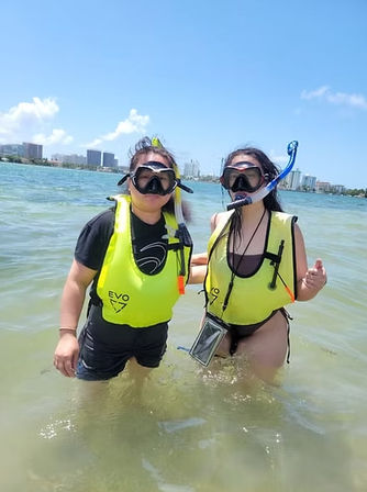 Two snorkelers in bright yellow life vests standing waist‑deep in clear tropical water near an urban shoreline with high‑rise buildings under a bright blue sky