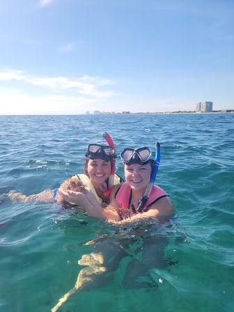 Two smiling snorkelers in masks, snorkels, and life vests floating in clear turquoise ocean water with a sandy coastline and beachfront buildings on the horizon under a sunny blue sky.