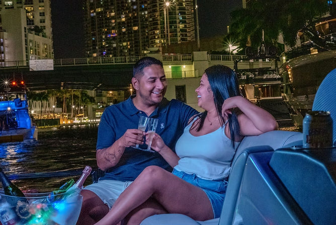 Smiling couple toasting with champagne on a yacht at night, city skyline and marina lights reflecting on the water