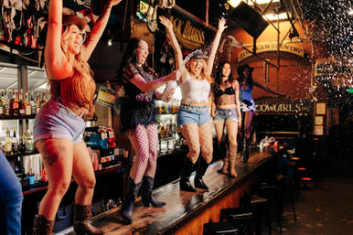 Five women in cowgirl outfits and cowboy boots dancing on a bar counter, cheering as one sprays champagne in a lively country bar nightlife scene.