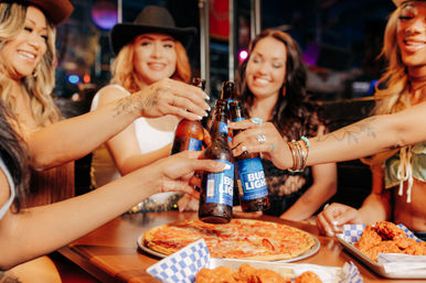 Friends toasting with bottled beers over pizza and chicken wings at a lively bar or pub during a night out.