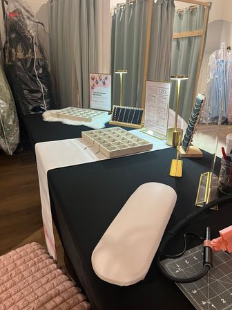 Pop-up jewelry display on a black table with necklace and charm trays, lit brass lamps and a price list in a boutique dressing room with hanging gowns and garment bags.