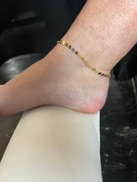 Close-up of a bare foot and ankle wearing a dainty gold chain anklet, foot resting on a white cushion against a dark background.