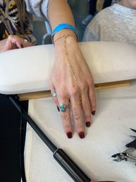 Close-up of a hand at a nail salon manicure on a white armrest — glossy burgundy nails, turquoise and silver rings, delicate gold hand-chain from wrist to finger, blue paper wristband and manicure tools nearby