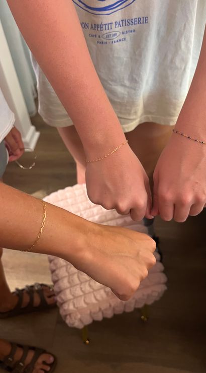 Three wrists wearing delicate gold chain and blue-beaded bracelets posed over a pink tufted stool on a hardwood floor in a casual indoor setting.