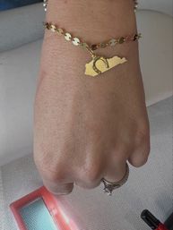 Close-up of a wrist wearing a gold disc-link bracelet with a Kentucky-shaped charm and small horseshoe; hand sports a diamond engagement ring with a pink compact mirror and nail polish nearby.