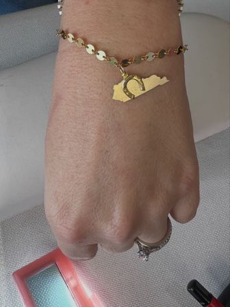 Close-up of a wrist wearing a gold disc-link bracelet with a Kentucky-shaped charm and small horseshoe; hand sports a diamond engagement ring with a pink compact mirror and nail polish nearby.
