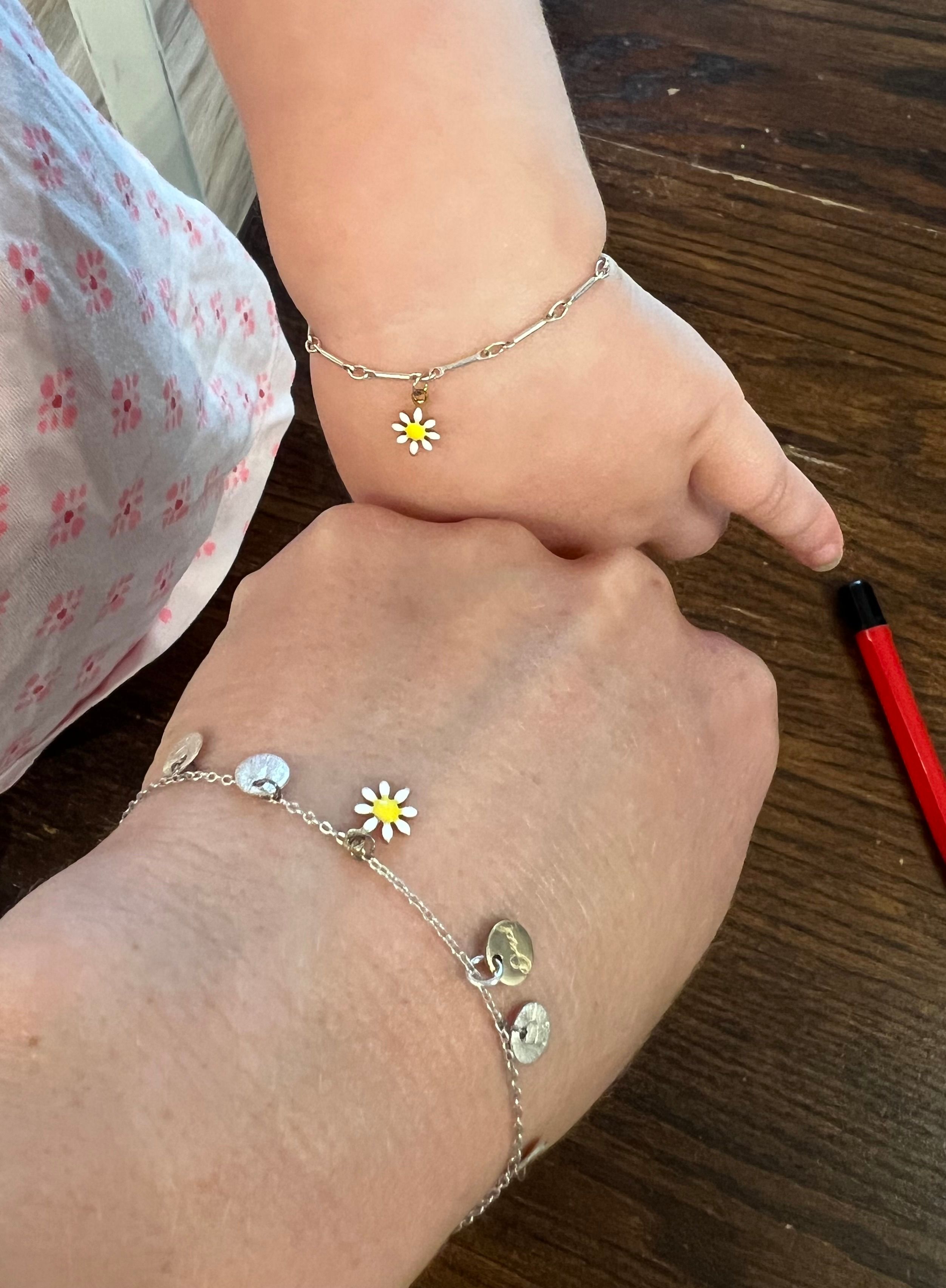Close-up of adult and child wrists wearing matching silver chain bracelets with small white-and-yellow daisy charms, resting on a dark wooden table with a red pen at the edge.